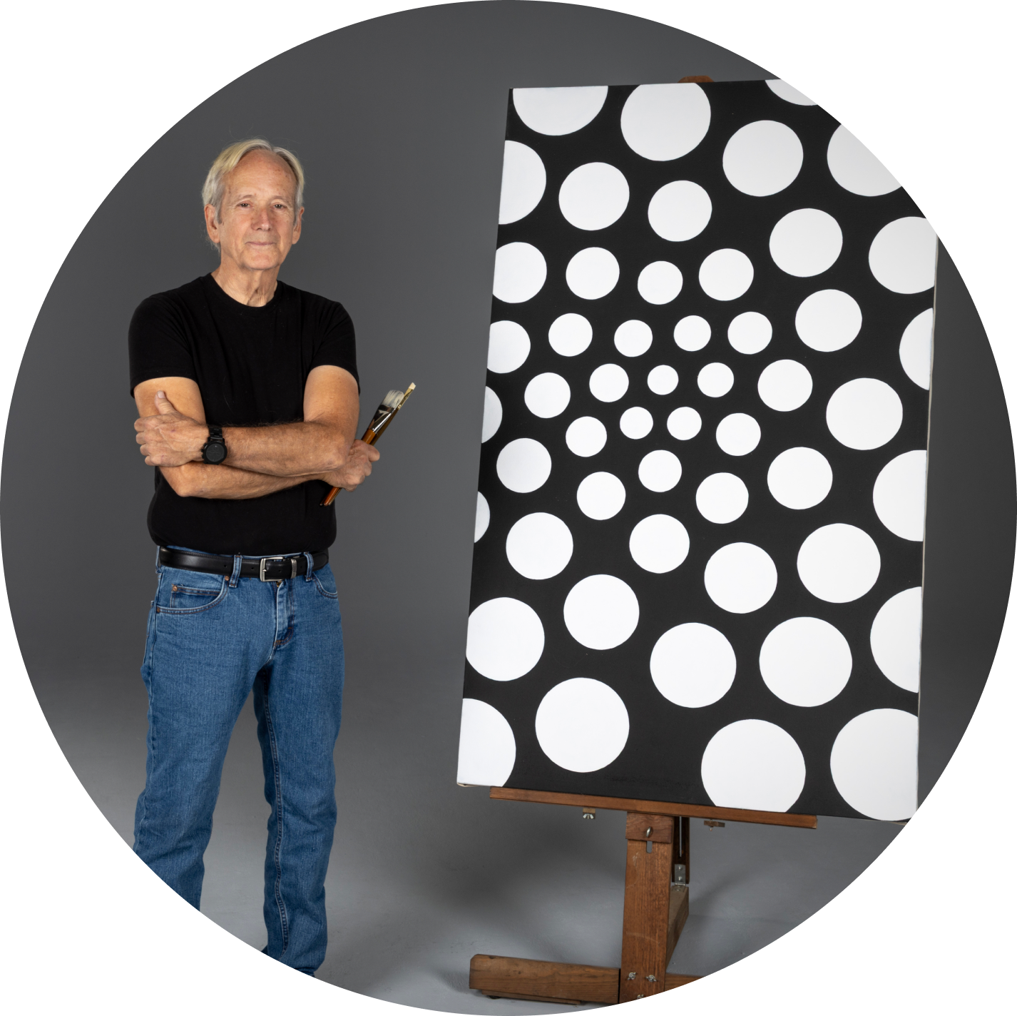 John Hopkins standing with arms crossed beside one of his dot paintings on an easel.